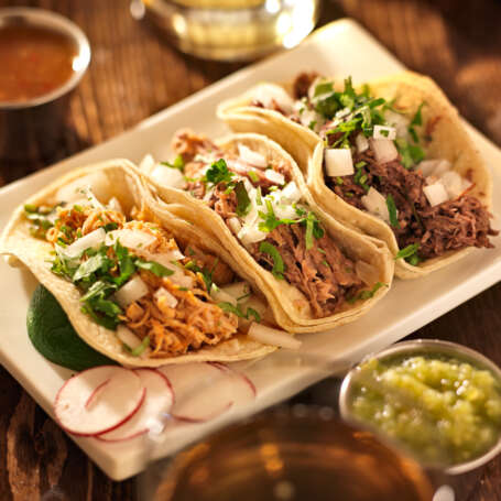 Three tacos on a white plate, served at a Mexican restaurant, showcasing vibrant toppings and fresh ingredients.